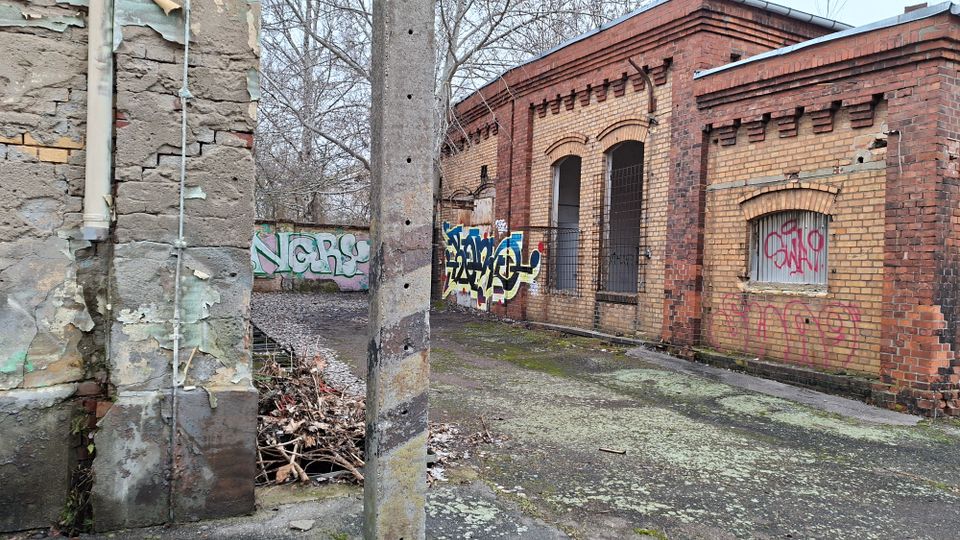 Verlassener Industriebau aus gelben und roten Ziegeln mit bogenförmigen Fenster- und Türöffnungen, teils vergittert und mit Graffiti besprüht. Davor liegt ein rissiger, moosiger Betonhof mit einem Betonpfosten, einer bröckelnden Wand und kahlen Bäumen im Hintergrund.