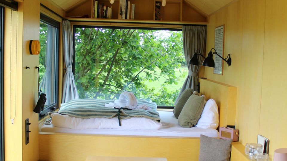 Innenansicht eines kleinen Tiny Houses mit holzverkleidetem Satteldach, eingebauter Schlafnische vor einem großen Fenster mit Blick ins Grüne und integrierten Regalen darüber. Vorne stehen ein kleiner Tisch, gepolsterte Sitzbänke und rechts eine kompakte Küchenzeile mit Kochfeld und Spüle.