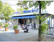 Außenbereich eines Cafés mit der Beschriftung „Café Winterfeld“ auf einem blauen Vordach. Vor dem Café stehen mehrere weiße Tische und Stühle, einige Gäste sitzen an den Tischen. Im Vordergrund sind bepflanzte Blumenkübel und ein Baum erkennbar.