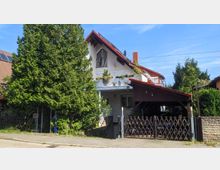 Zweistöckiges Einfamilienhaus mit hellen Wänden und einem roten Satteldach, umgeben von Bäumen und Sträuchern. Vor dem Haus befindet sich ein überdachter Carport mit Holzzaun, links ein großes grünes Tor mit Bogen.