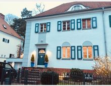Zweistöckiges Wohnhaus mit weißer Putzfassade, blauen Fensterläden und Satteldach aus roten Ziegeln. Vor dem Haus befinden sich eine Stufenanlage, dekorative Pflanzen in blauen Töpfen und ein schwarzer Zaun; links ist ein angrenzendes Haus mit ähnlicher Architektur sichtbar.