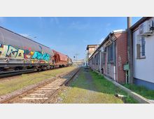 Ein Industriegebiet mit einer Bahngleisanlage, auf der ein Güterzug mit Graffiti steht. Rechts befindet sich ein langes Backsteingebäude mit vergitterten Fenstern und Klimaanlagen, daneben ein schmaler Grasstreifen und ein Fußweg.