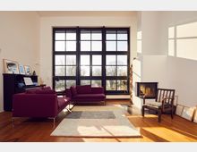 A modern living room with large floor-to-ceiling windows divided into black-framed panes, allowing natural light to fill the space. The room features maroon sofas, a piano with framed artwork on top, a geometric area rug on a wooden floor, and a fireplace with stacked firewood nearby.