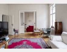 Helles Wohnzimmer mit Parkettboden, einem großen bunten Teppich und einer weißen Couch. Im Hintergrund führen weiße Flügeltüren zu einem weiteren Raum mit einem roten Sofa, und auf der rechten Seite des Raumes stehen ein kleiner Tisch, ein Korbstuhl und ein Holzschrank.