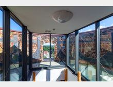 Innenansicht eines modernen Gebäudes mit großen Glasfenstern, die einen Blick auf eine historische Ziegelmauer mit einer offenen Bogenöffnung freigeben. Im Hintergrund sind grüne Landschaften und Dächer von Häusern sichtbar.