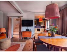 Wohnzimmer mit offenem Essbereich und Küche. Holzmöbel und -boden dominieren den Raum; auffällige Elemente sind eine hölzerne Treppe, pinkfarbene Lampenschirme, Vorhänge sowie ein Esstisch mit Vase voller Blumen. Im Hintergrund befindet sich eine Küche mit cremefarbenen Schränken.