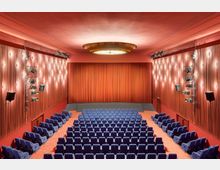 Innenraum eines klassischen Kinos mit roten Vorhängen an der Bühne und den Wänden, blauer Bestuhlung in Reihen sowie einer zentralen Deckenleuchte. Die Wände sind mit warmen Leuchten und Beleuchtungselementen verziert.