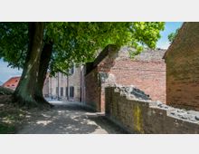 Ein schmaler Weg verläuft zwischen einer Backsteinmauer mit teilweise beschädigten Abschnitten und einem historischen Gebäude mit Fenstern und Gitterstäben. Links steht ein großer Baum mit dichtem Blattwerk, das Schatten auf den Weg wirft, und im Hintergrund sind Häuser mit roten Ziegeldächern zu sehen.