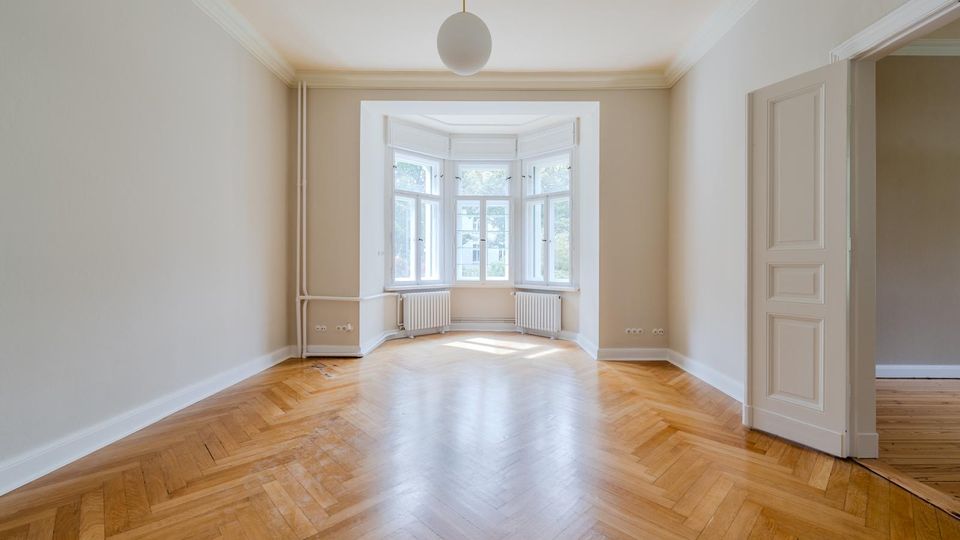 Heller, leerer Altbau-Wohnraum mit Fischgrätparkett, hohen Decken mit Stuck und einer zentralen Pendelleuchte. Am Ende des Raums befindet sich ein großer Erker mit mehreren Fenstern und Heizkörpern darunter; rechts ist eine offene Tür zu einem angrenzenden Zimmer zu sehen.