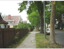 Wohnstraße mit gepflastertem Gehweg und angrenzendem Rasenstreifen. Rechts stehen Bäume und parkende Autos entlang der Straße, links ein Holzzaun und Wohnhäuser mit roten Ziegeldächern und Gärten im Hintergrund.