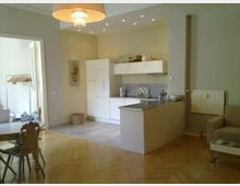 Offene Wohnküche mit modernem Design, bestehend aus weißen Einbauschränken, einer kleinen Kücheninsel mit Arbeitsfläche aus Edelstahl und Holzboden in Fischgrätmuster. Im Hintergrund durch eine Tür ein separater Raum mit Kleiderstange; im Wohnbereich ein Esstisch mit Holzstühlen und ein Sessel.