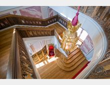 Holzverkleidetes Treppenhaus mit aufwendig geschnitztem Geländer und hölzernem Boden, einem roten Thronstuhl auf einem Podest sowie einer goldenen, verzierten Laterne, die von der Decke hängt. Die Wände sind teilweise mit rosa marmorierten Paneelen gestaltet.