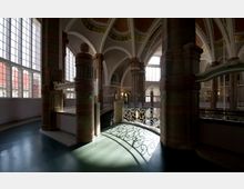 Innenansicht eines historischen Gebäudes mit hohen Säulen aus Sandstein, großen Fensterflächen, die Tageslicht hereinlassen, und dekorativen Gewölbedecken. Im Vordergrund eine geschwungene Brüstung mit verschnörkelten Mustern, die einen Schatten auf den Boden wirft.