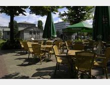 Außenbereich eines Cafés mit zahlreichen Holztischen und Stühlen sowie großen grünen Sonnenschirmen, umgeben von Bäumen und Gebäuden im Hintergrund. Die Szene befindet sich auf einem gepflasterten Platz bei Tageslicht.