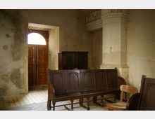 Innenraum eines alten Gebäudes mit abgenutzten Wänden und einer leicht beschädigten, hell gestrichenen Säule. Im Vordergrund befinden sich dunkle, hölzerne Kirchenbänke und ein einzelner Stuhl, im Hintergrund ist eine Holz-Doppeltür mit einem halbkreisförmigen Fenster zu sehen.