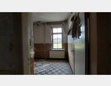 Alte, verlassene Küche mit abgenutzten Wänden, brüchigen Decken und einem schmutzigen Schachbrett-Boden. Ein Fenster mit beschädigtem Rahmen zeigt eine Wiese und Gebäude im Hintergrund.