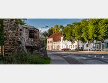 Eine Straßenkreuzung mit einem mittelalterlichen Mauerrest aus Natursteinen und Ziegeln auf der linken Seite. Im Hintergrund stehen moderne Wohnhäuser mit weißen Fassaden und roten Dächern, daneben Bäume entlang des Gehwegs.