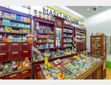 Historische Drogerie mit dunkelroten Holzregalen, gefüllt mit diversen Produkten wie Seifen, Verpackungen und Flaschen, sowie einer Glastheke mit weiteren ausgestellten Artikeln. Im Hintergrund ist ein Schild mit der Aufschrift „Markt Drogerie“ und mehrere dekorative Vorratsgläser zu sehen.