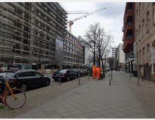 Straßenszene in einer urbanen Umgebung mit einem im Bau befindlichen Gebäudekomplex auf der linken Seite, eingerüstet und von Baukränen umgeben. Auf der rechten Seite befinden sich Wohngebäude, teils mit roten Fassaden, daneben Gehwege mit Fußgängern und orange gekleideten Arbeitern.