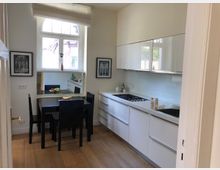 Moderne Küche mit weißen Hochglanzschränken, integriertem Kochfeld und Holzfußboden. Ein Esstisch mit schwarzen Stühlen steht vor einem Fenster mit gewölbtem oberen Rahmen.