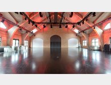 Großer Veranstaltungsraum mit einer hohen Decke, Betonträgern und Ziegelwänden, ergänzt durch große Fenster und eine zentrale, schwarze Tür. Eine Lichtinstallation hängt unter der Decke, während der Raum mit dunklem, reflektierendem Boden und weißen Tischen entlang der Seite ausgestattet ist.