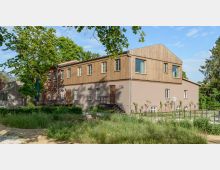 Zweistöckiges Gebäude mit einer Fassade aus Holz und Putz, umgeben von Bäumen und Grünflächen. Das Gebäude verfügt über große Fenster im oberen Stockwerk, Treppen vor den Eingangstüren und liegt in einer parkähnlichen Umgebung bei klarem Himmel.