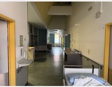 Ein Krankenhausflur mit grauem Boden und gelb umrandeten Türen. Auf der rechten Seite steht ein leeres Pflegebett, während links eine Handdesinfektionsstation und ein Wandspiegel sichtbar sind; das Ende des Flurs ist durch ein Fenster beleuchtet.