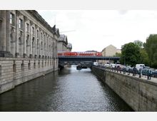 Historisches Gebäude mit Steinfassade am Wasserkanal, über dem auf einer Brücke ein roter Zug fährt; rechts führt eine Straße mit parkenden Autos und Gehweg entlang des Kanals.