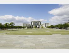 Moderner Regierungsbau mit symmetrischem, kubischem Design, umgeben von offenen Grünflächen und gepflasterten Gehwegen. Im Vordergrund eine große, gestreifte Rasenfläche, eingerahmt von Baumreihen; im Hintergrund ein blauer Himmel mit weißen Wolken.