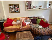 Ein gemütliches Wohnzimmer mit einer bunt gemusterten Eckcouch, die mit roten, grünen und gemusterten Kissen dekoriert ist. Eine Person liegt auf der Couch und liest ein Comic-Heft; an der Wand hängt eine Pinnwand mit Fotos und darüber ein großer Spiegel, der den Raum reflektiert.