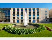Moderne Gebäudefassade mit großen quadratischen Fenstern im Hintergrund, davor ein runder Springbrunnen, umgeben von grünen Pflanzen und gepflegtem Rasen. Der Himmel ist klar und blau.
