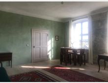 Ein heller Raum mit grünen, verputzten Wänden, einer Holzdecke und Holzdielenboden. Im Raum stehen ein Tisch mit vier Stühlen vor einem großen Fenster mit weißen Vorhängen; daneben sind Teppiche, ein Beistelltisch und ein Sideboard sichtbar.