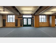 Großer Innenraum mit Backsteinwänden, zwei großen Fenstern und einer zentralen grünen Glastür mit Kreuzverstrebungen. Der Boden ist mit grauem Teppich ausgelegt, und es gibt sichtbare Deckenbalken sowie hölzerne und metallene Trennwände.