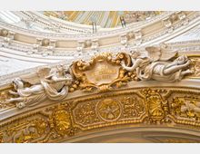 Detailaufnahme eines barocken Kirchenelements mit reich verzierter Architektur, goldenen Ornamenten und Figuren von Engeln, die ein Schriftfeld mit der Aufschrift „Lasset euch versöhnen mit Gott“ einrahmen. Im unteren Bereich ist ein Relief mit einer Taube, umgeben von floralen Mustern, zu sehen.