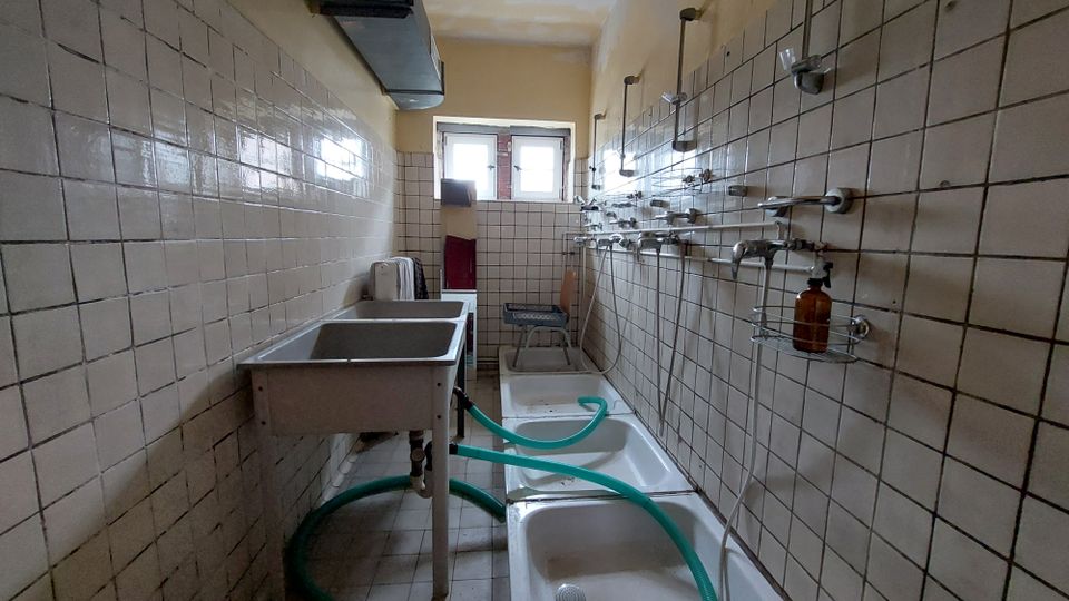 Ein schmaler Duschraum mit beigen gefliesten Wänden und mehreren Duscharmaturen auf der rechten Seite. Links befinden sich ein Edelstahlwaschbecken und dahinter ein Fenster, das etwas Licht in den Raum bringt.