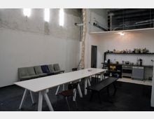 Industriell gestalteter Raum mit einer einfachen Küche auf der rechten Seite, ausgestattet mit Herd, Spüle und Regalböden, sowie einem langen weißen Tisch mit Stühlen und einer Bank im Vordergrund. Links an der Wand stehen gepolsterte Stühle unter rauen, unverputzten Wänden mit sichtbaren Kabeln und Leuchten.