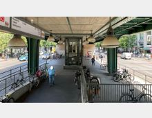 Überdachter U-Bahn-Zugangsbereich mit grüner Metallkonstruktion, großen Hängelampen und einem gläsernen Aufzug in der Mitte. Auf beiden Seiten verlaufen Gehwege mit geparkten Fahrrädern, daneben eine belebte Straße mit Autos und Fußgängern.