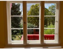 Blick durch ein weiß gerahmtes Fenster auf einen kleinen Garten mit grünen Bäumen und Sträuchern. Im Vordergrund befindet sich ein rotes Geländer, und im Hintergrund sind ein Teil eines Hauses und ein blauer Himmel sichtbar.