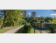 Ein Park mit einem gepflasterten Weg, umgeben von Bäumen und Sträuchern. Im Hintergrund ragen drei mehrstöckige rote Wohngebäude über die Baumkronen hinaus, rechts stehen flache Garagen.