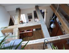 Innenansicht eines mehrstöckigen Wohnraums mit sichtbaren, groben Holzbalken als Zwischendecke. Im unteren Bereich sind eine Holzküche, eine weiße Tür sowie dekorative Elemente zu erkennen, während Pflanzen und eine Hängelampe den Raum ergänzen.