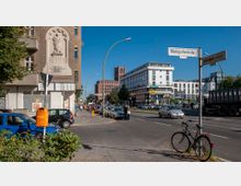 Straßenszene mit mehrspuriger Straße und Gehwegen, flankiert von modernen und älteren Gebäuden, darunter ein Backsteingebäude mit Uhrturm und ein weißer moderner Bau. Im Vordergrund ein Fahrrad, parkende Autos und ein orangefarbener Mülleimer; Straßenschilder zeigen „Markgrafenstraße“ und „Mariendorfer Damm“.