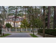 Zweistöckige Villa mit gelber Fassade und rotbraunem Ziegeldach, umgeben von einem Garten mit vielen Bäumen. Ein weißer Zaun und eine ruhige, baumgesäumte Straße grenzen das Grundstück ab.