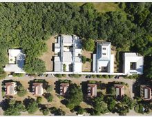 Luftaufnahme einer Anlage mit mehreren rechteckigen Gebäuden in heller Farbe, umgeben von dichtem Wald. Im unteren Bereich sind kleinere Wohnhäuser mit roten Dächern zu sehen, dazwischen geordnete Grünflächen und Parkplätze.