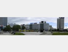 Weitläufiger Stadtplatz mit Hochhäusern und moderner Bebauung im Hintergrund, darunter markante Werbeschilder. Im Vordergrund sind grüne Rasenflächen, Gehwege, Passanten und eine gelbe Straßenbahn zu sehen.