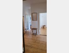 Ein heller Flur mit Holzdielenboden, einer kleinen Holzkonsole an der Wand und einem rechteckigen Spiegel darüber. Im Hintergrund sind zwei geöffnete Türen, die in ein Badezimmer und ein weiteres Zimmer mit Korb und Kleiderhaken führen.