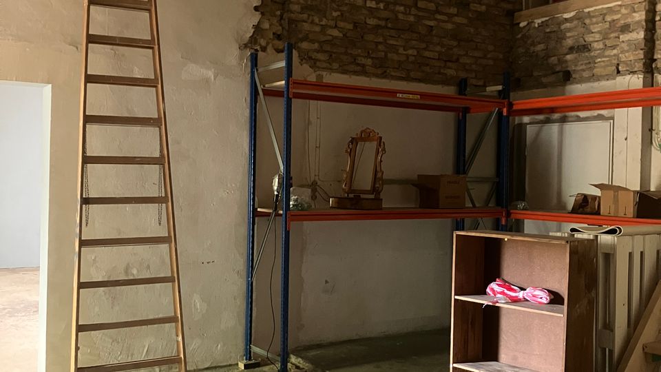 Interior of a warehouse or storage room with a concrete floor, exposed brick and plaster walls, and a high sloped ceiling. A tall wooden ladder leans against the wall beside metal shelving with boxes, with a small mezzanine railing and scattered furniture pieces in the foreground.