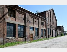 Ziegelsteinfassade eines alten, industriellen Lagergebäudes mit großen, rechteckigen Fenstern und einer Holztür, umgeben von einem gepflasterten Weg und etwas Bewuchs. Der Himmel ist klar und blau, was auf eine Außenaufnahme bei Tageslicht hinweist.