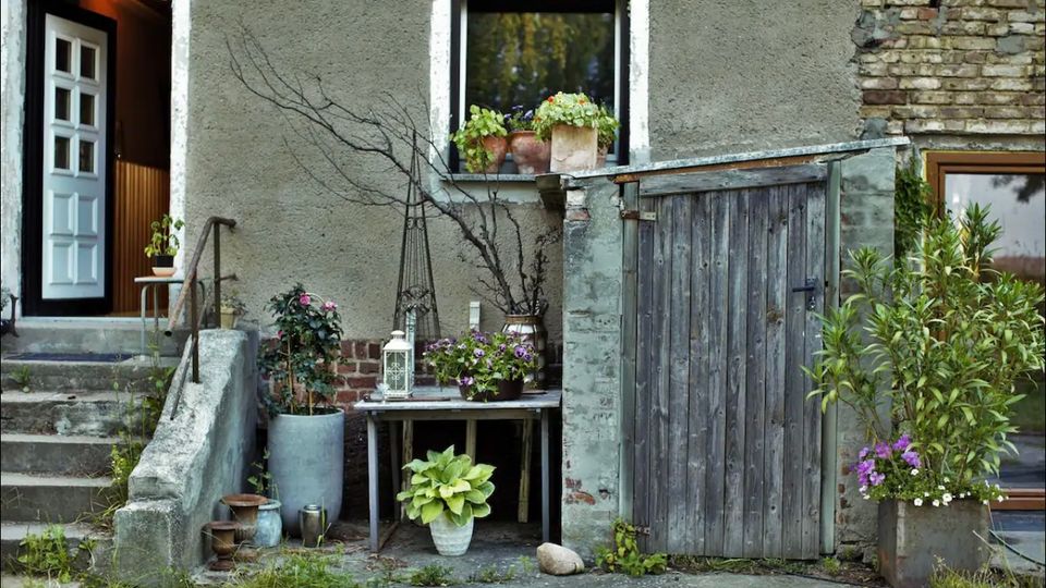Eine Hausfassade mit einer offenen weißen Tür auf der linken Seite, mehreren blühenden Pflanzen in Töpfen vor einem Fenster, sowie einem alten Holztisch und einem verwitterten Holztor auf der rechten Seite. Die Umgebung wirkt rustikal, mit sichtbaren Putz- und Backsteinflächen.