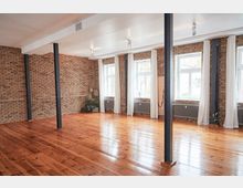 Heller Raum mit Holzboden, Backsteinwänden und großen Fenstern, die mit weißen Vorhängen dekoriert sind. Mehrere schwarze Metallstützen unterteilen den Raum, der von einer schlichten weißen Decke abgeschlossen wird.