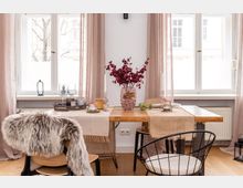 Esszimmer mit einem Holztisch, darauf Tischläufer, Kerzen, Dekoobjekte und eine Vase mit roten Zweigen. Im Hintergrund helle Vorhänge vor großen Fenstern, ein Heizkörper darunter, und Stühle mit unterschiedlichen Sitzkissen um den Tisch.